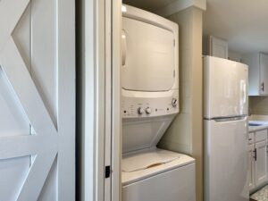 Stacked washer and dryer next to a refrigerator in a small Skipping Stone Lane laundry area with white cabinets.