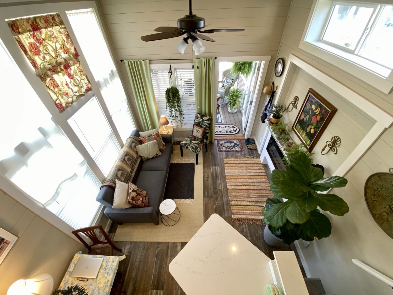 Bright cozy living room on Skipping Stone Lane with large windows, plants, colorful rugs, and a gray sofa seen from above.