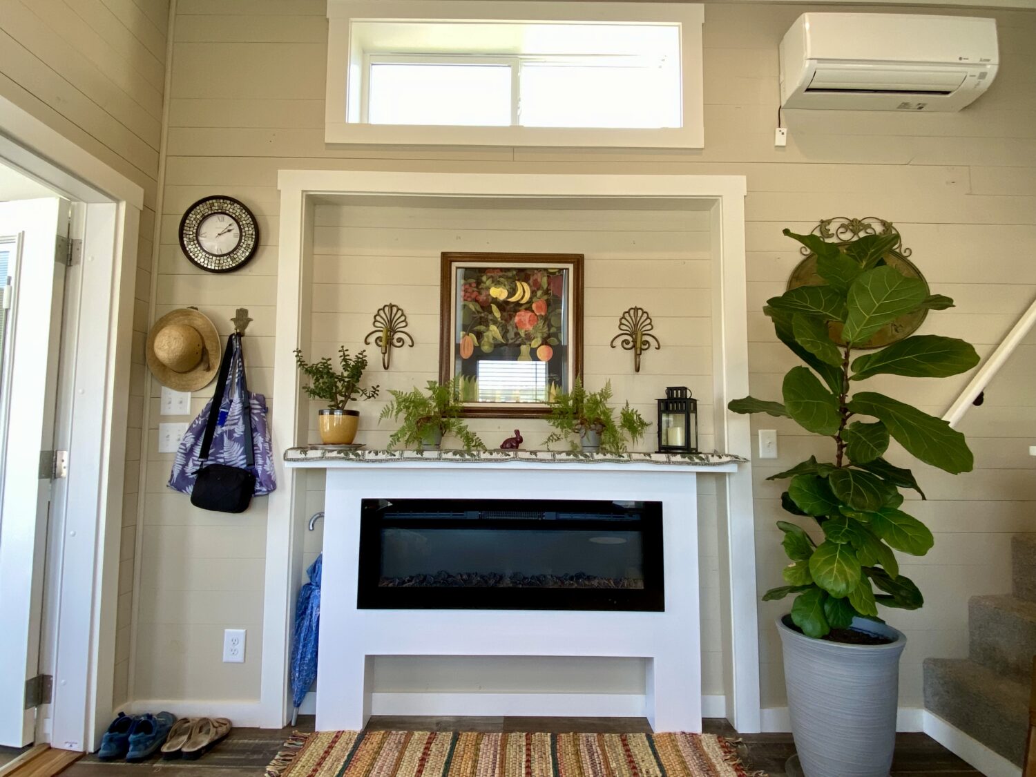 A cozy entryway at 43 Skipping Stone Lane with a fireplace, potted plant, wall clock, hats, and a striped rug.