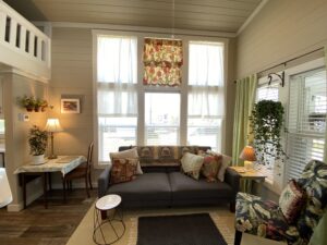 Bright living room on Skipping Stone Lane with a gray sofa, floral pillows, and large windows with sheer curtains.