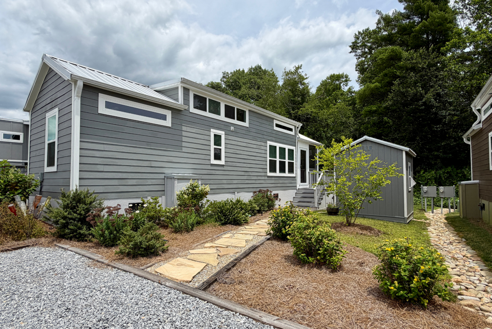 A modern tiny house with gray siding and a stone walkway on Mount Meadow Lane, set beneath a cloudy sky.