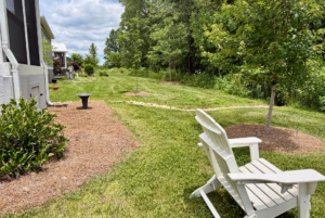 White chair on lawn near trees and mulch in a Mount Meadow Lane backyard, path and house on left, cloudy sky above.