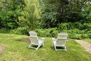 Two white Adirondack chairs on a green lawn along Mount Meadow Lane, facing dense trees and greenery on a sunny day.