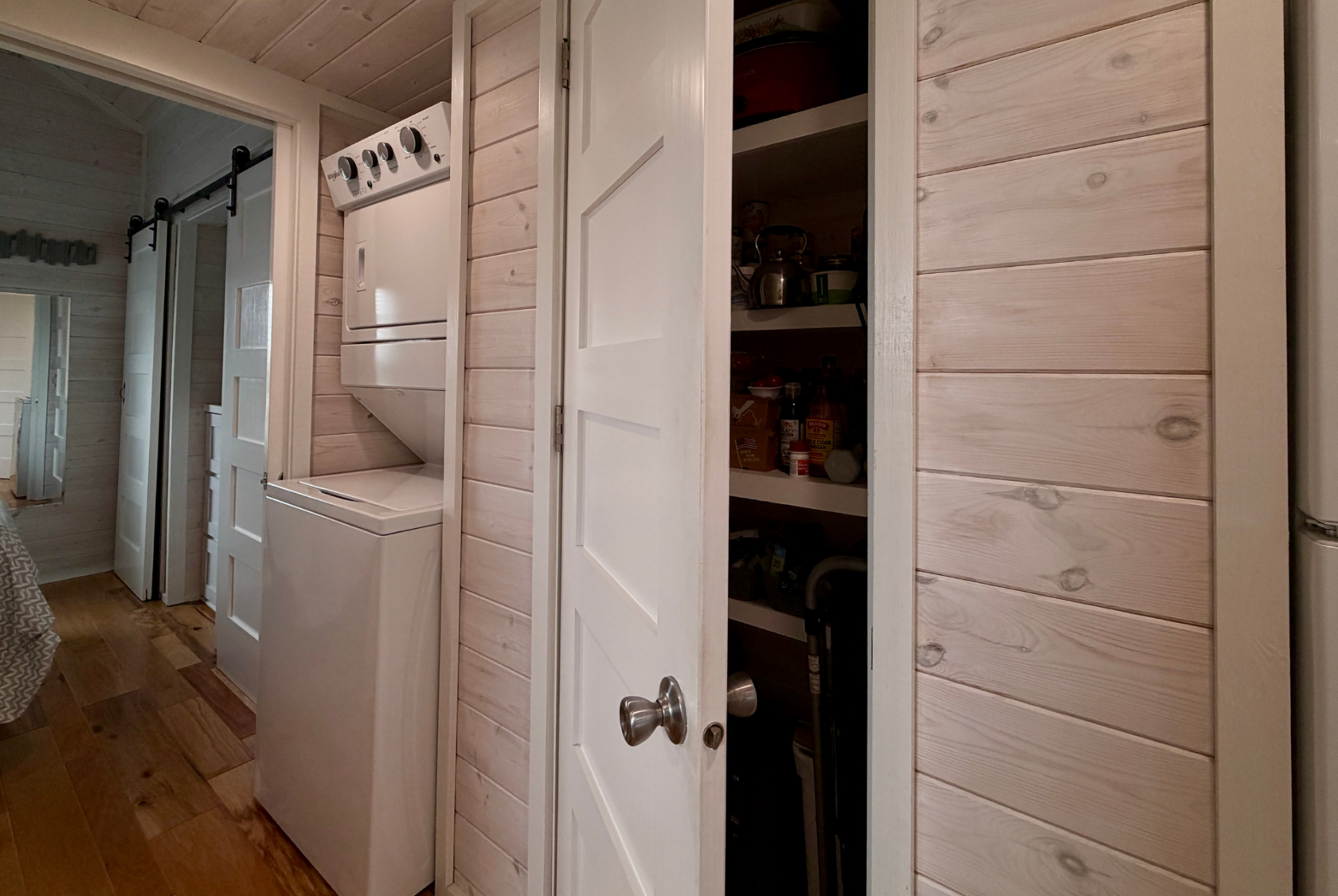 A small Mount Meadow Lane laundry area with a stacked washer-dryer sits beside an open pantry door in a wooden-paneled room.