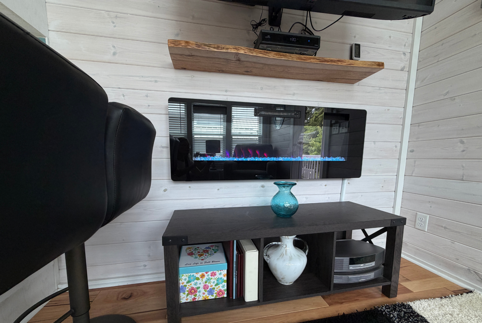 Electric fireplace with blue flames on a white wall at Mount Meadow Lane, black chair, and dark table with decor below.