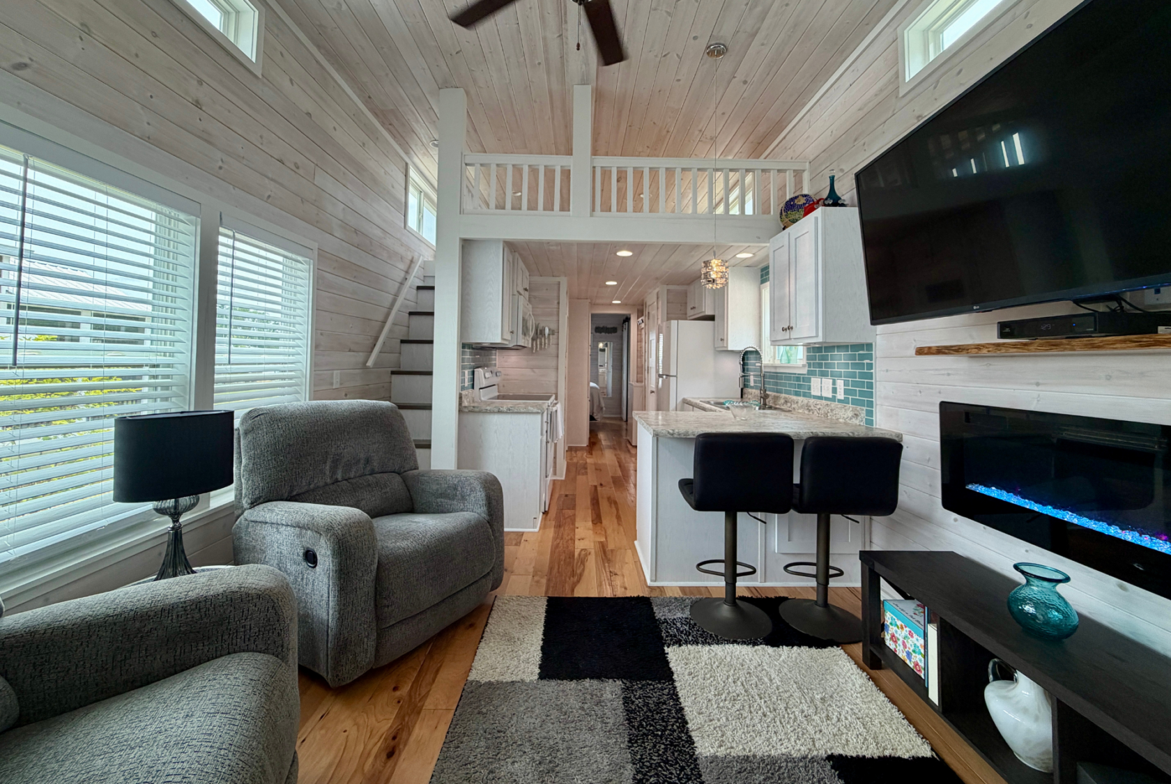 Modern Mount Meadow Lane tiny house interior with gray armchair, kitchen, loft, bar stools, TV, and large windows.
