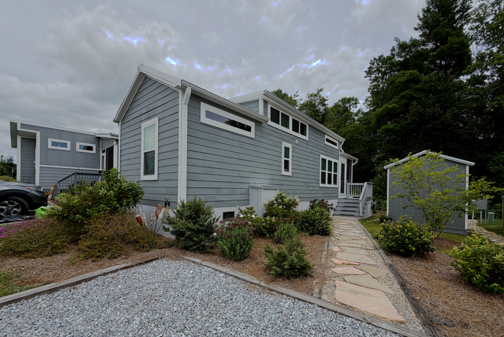 A modern gray tiny house with white trim sits on Mount Meadow Lane, surrounded by plants and a stone path.