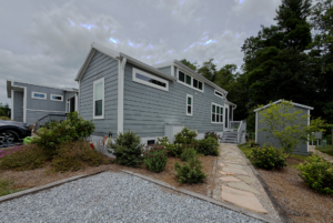 A modern gray tiny house with white trim sits on Mount Meadow Lane, surrounded by plants and a stone path.