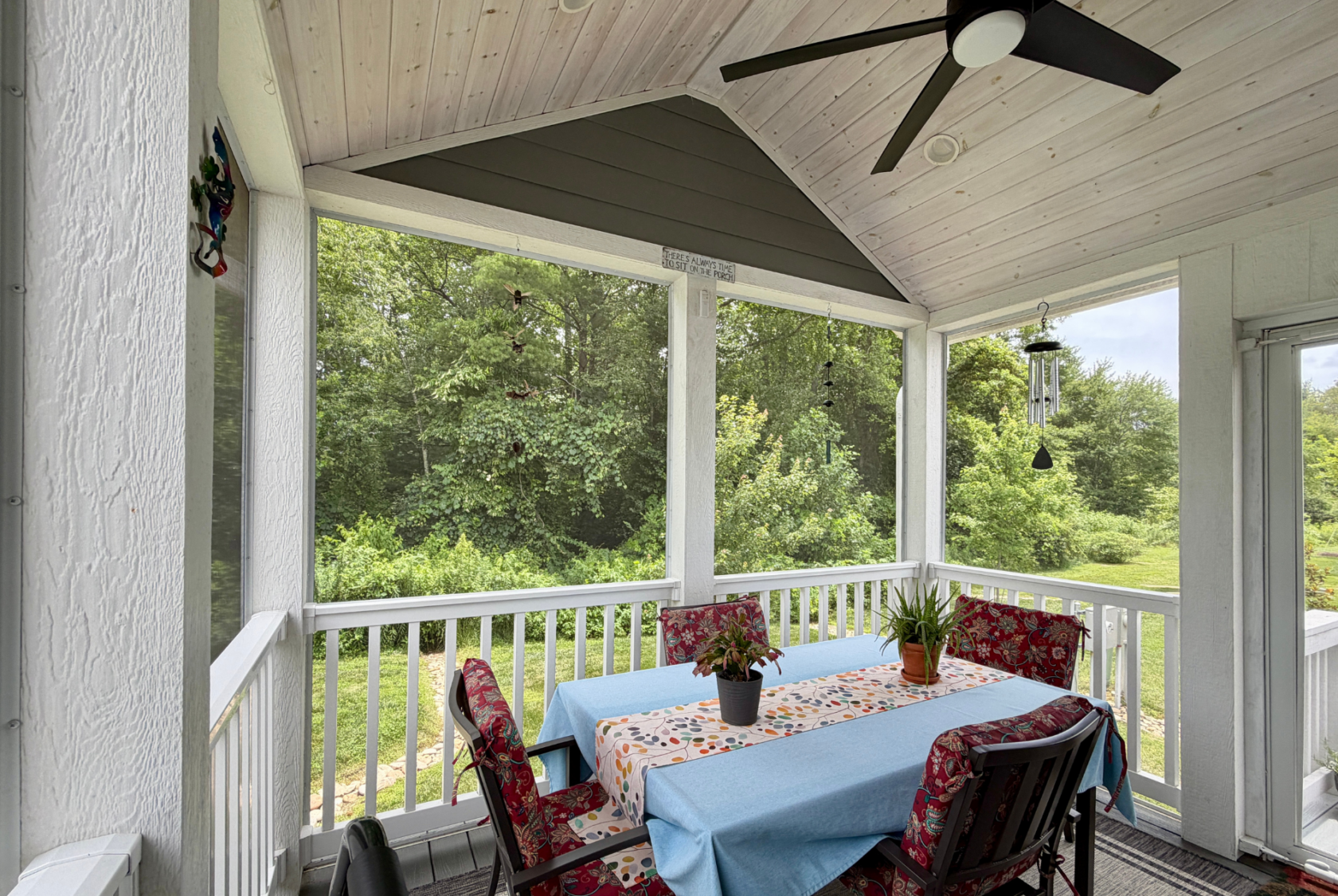 Screened porch on Mount Meadow Lane with a table, chairs, potted plants, and a ceiling fan, overlooking green trees.