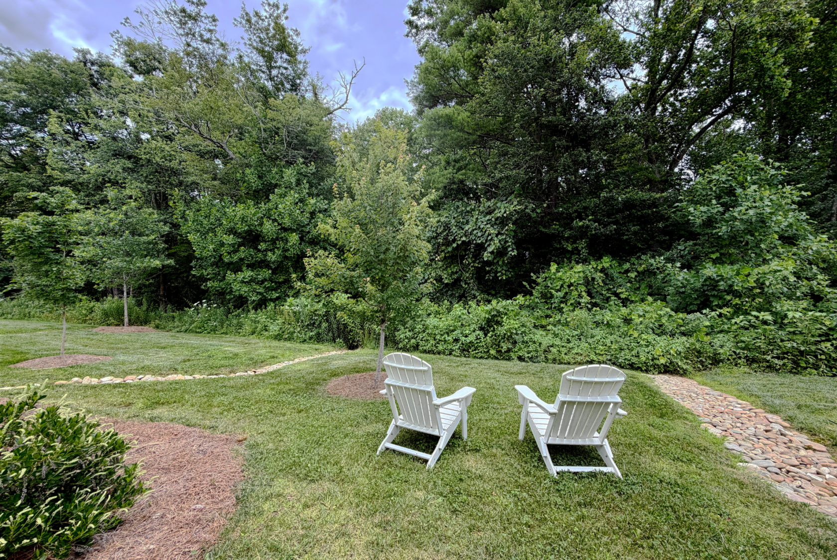 Two white Adirondack chairs overlook a lush forest on Mount Meadow Lane, with stone paths winding through the grass.