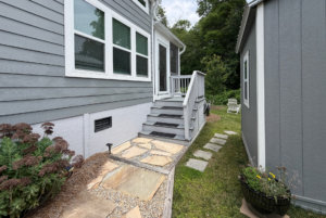 Stone path leads to porch of gray house on Mount Meadow Lane, with plants and lawn beside a gray shed.
