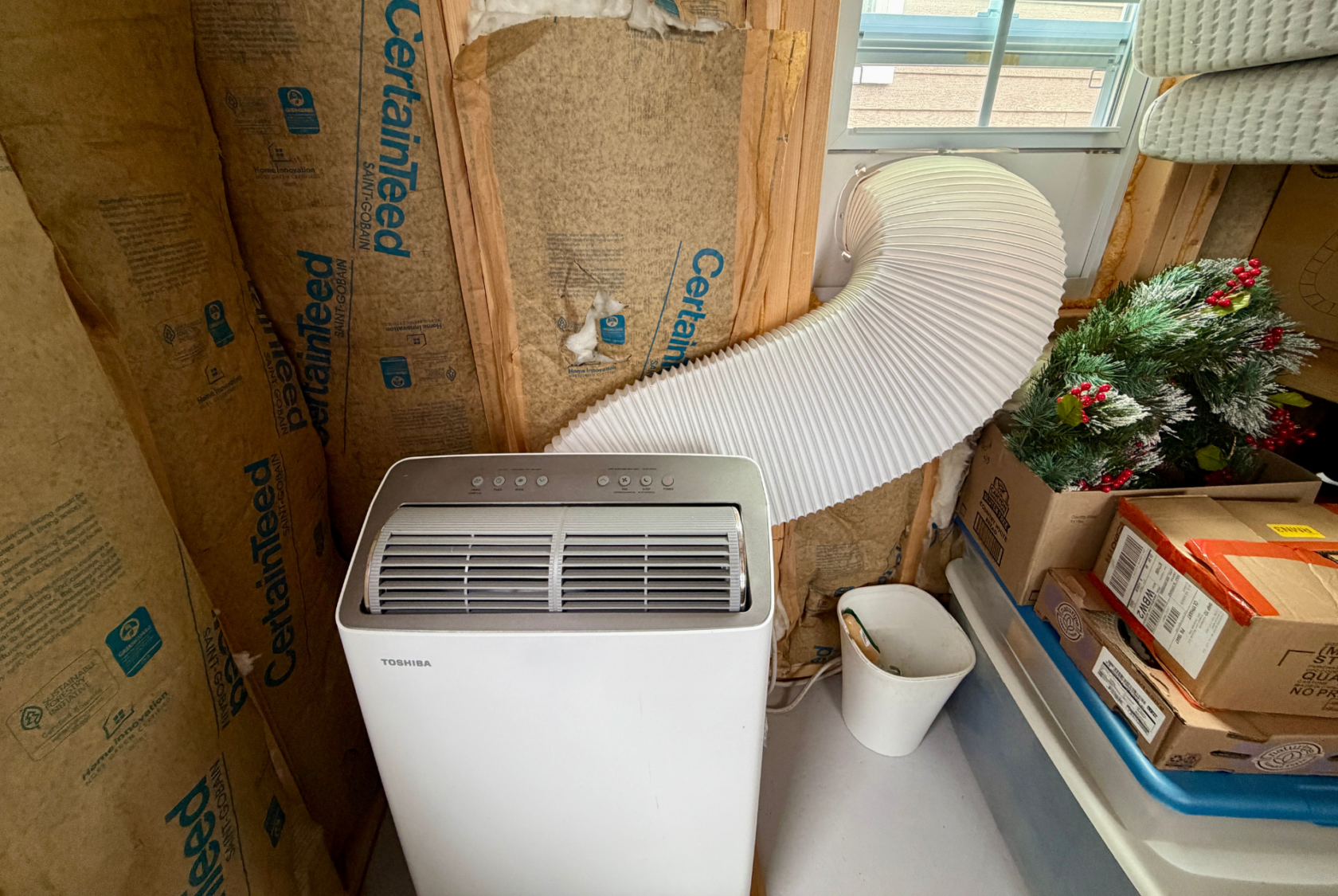 Portable air conditioner vented out a window in an unfinished Mount Meadow Lane room with boxes and holiday decor nearby.