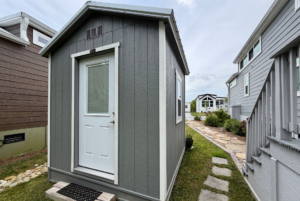 A small gray shed with a white door sits by a stone path on Mount Meadow Lane, near larger houses on a cloudy day.