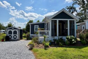 A small blue house on 103 Meandering Lane with a porch, garden, gravel driveway, and shed shines on a sunny day.