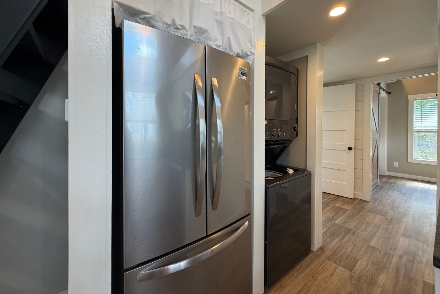 Stainless steel fridge and stacked washer dryer accent 103 Meandering Lane’s modern hallway with wood floors, white walls.