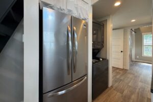 Stainless steel fridge and stacked washer dryer accent 103 Meandering Lane’s modern hallway with wood floors, white walls.
