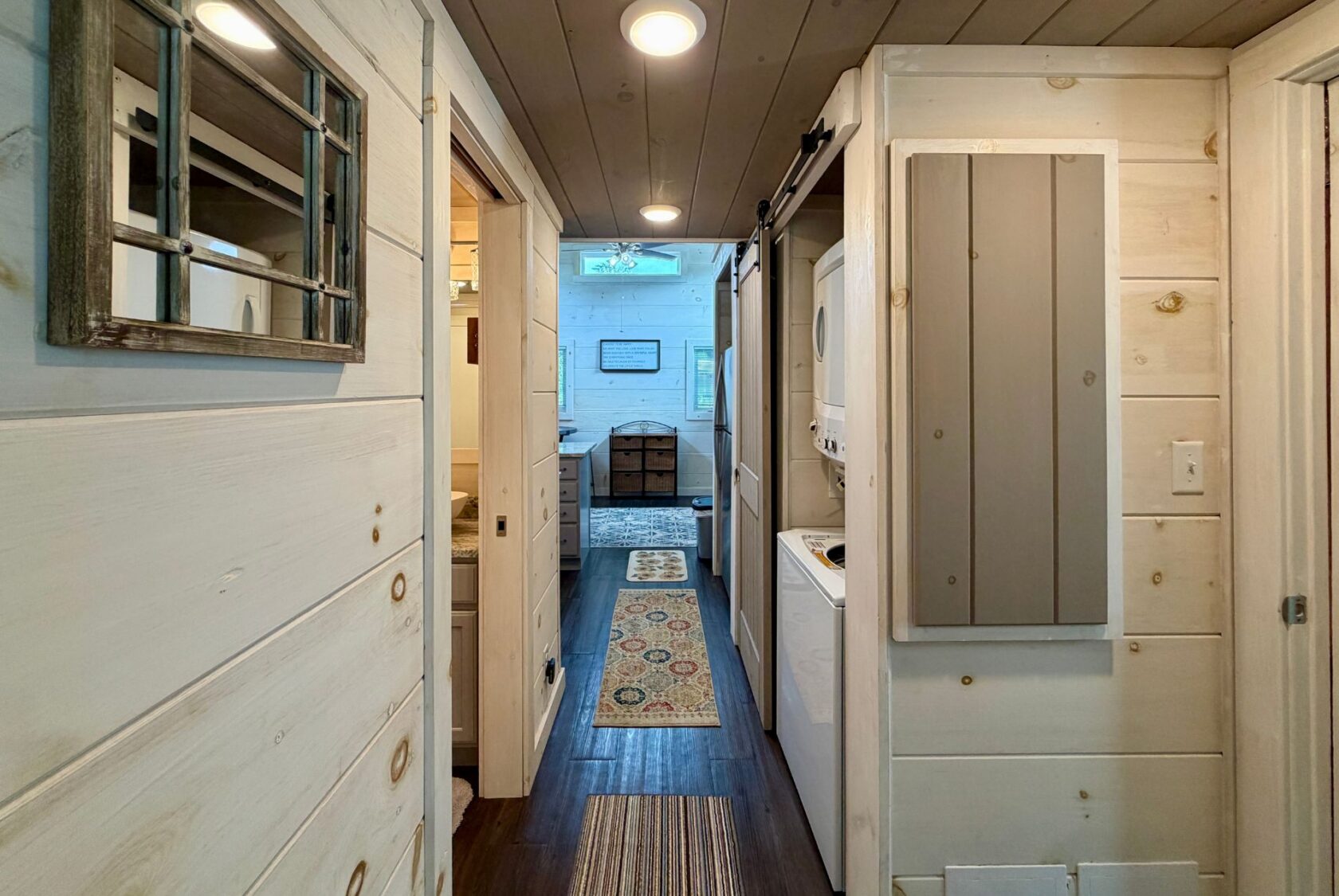 Narrow Highland Hill Lane hallway with white wooden walls, washer and dryer, leading to a cozy room with a rug.