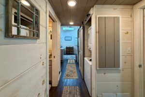 Narrow Highland Hill Lane hallway with white wooden walls, washer and dryer, leading to a cozy room with a rug.