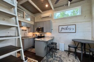 Modern Highland Hill Lane tiny kitchen with gray cabinets, marble island, “gather” sign, and loft ladder on the left.