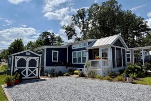A small blue house at 103 Meandering Lane with a white porch sits beside a gravel driveway under a sunny sky.