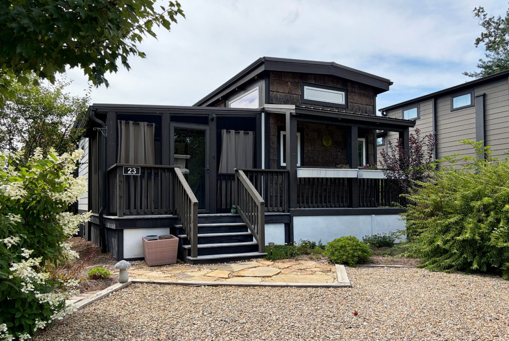 A small, dark house with a porch, gravel yard, plants, and steps sits on Highland Hill Lane; number 23 is by the door.
