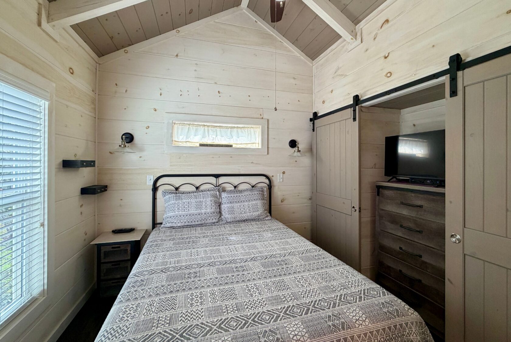 Cozy Highland Hill Lane bedroom with patterned bedding, wood walls, a dresser, TV, and barn-style closet doors.