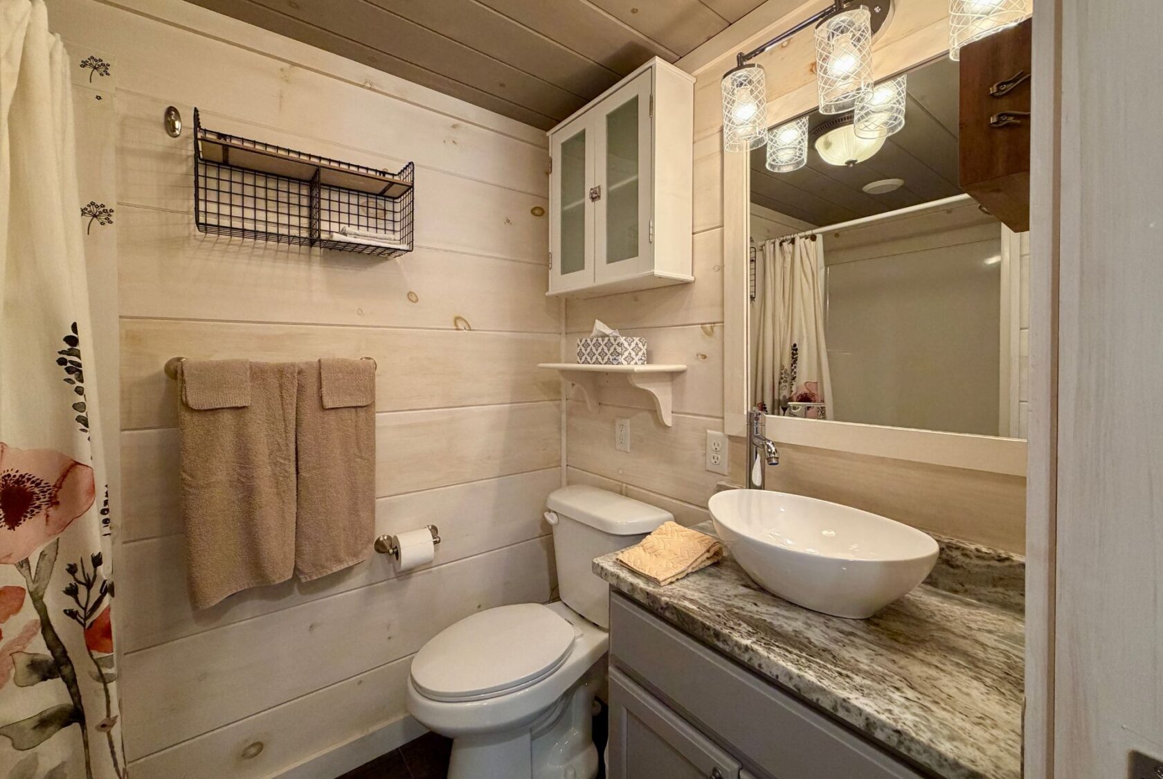 Cozy Highland Hill Lane bathroom with wood walls, vessel sink, towel rack, wall shelves, and mirrored vanity with lights.