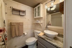Cozy Highland Hill Lane bathroom with wood walls, vessel sink, towel rack, wall shelves, and mirrored vanity with lights.