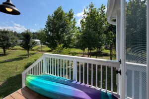 A colorful kayak rests on a porch at 103 Meandering Lane, overlooking a sunny yard with green trees and grass.