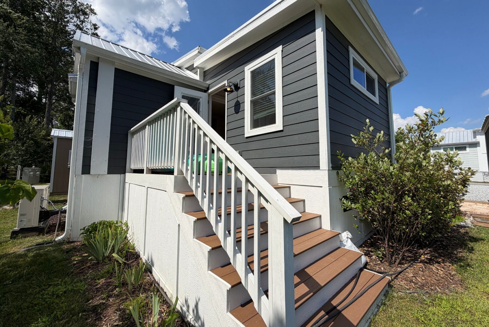 A small modern house at 103 Meandering Lane with gray siding, white trim, and a front staircase amid lush plants.