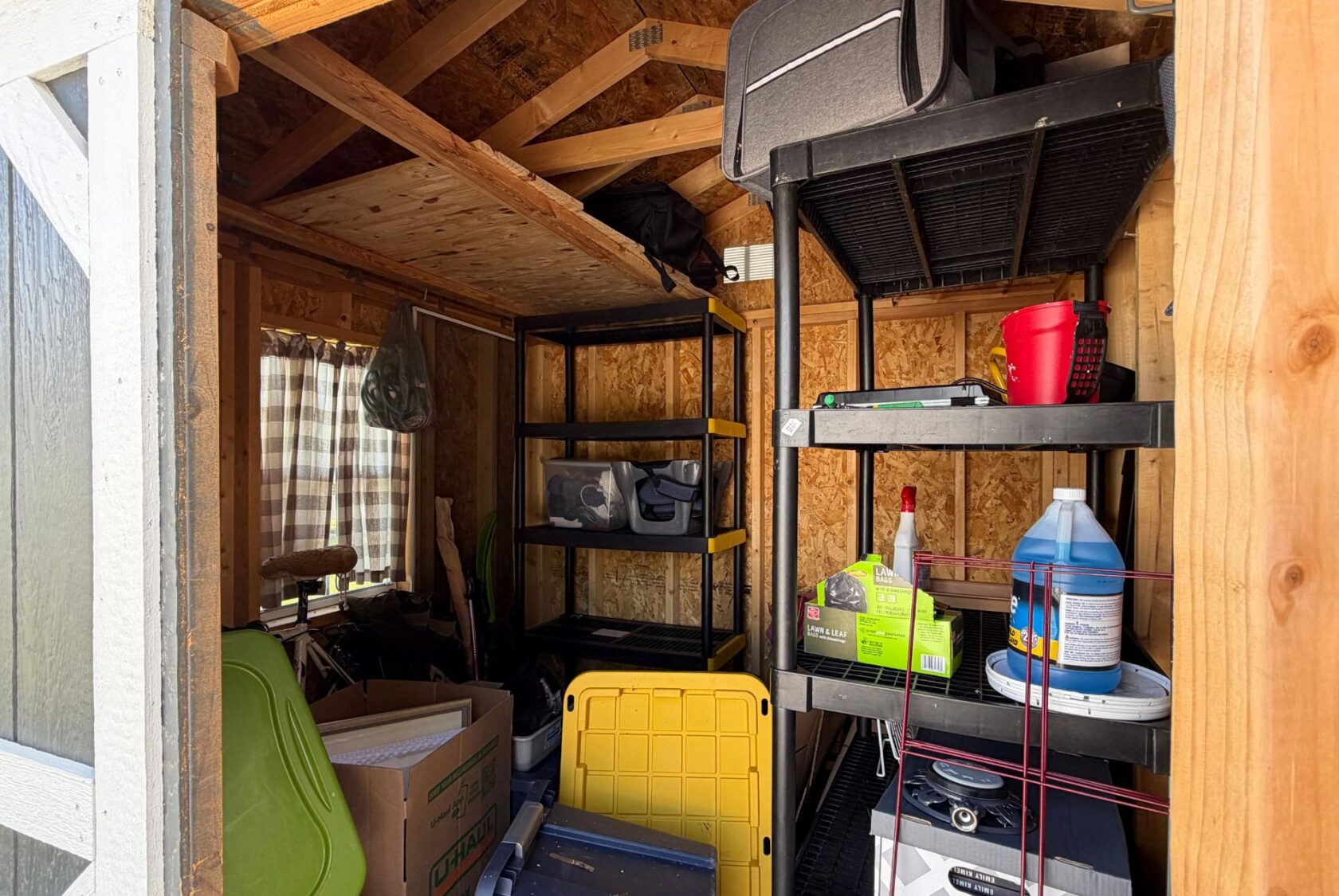 A cluttered shed at 103 Meandering Lane with shelves, boxes, cleaning supplies, and storage bins against wooden walls.