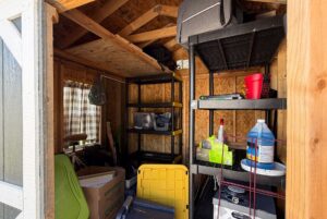 A cluttered shed at 103 Meandering Lane with shelves, boxes, cleaning supplies, and storage bins against wooden walls.
