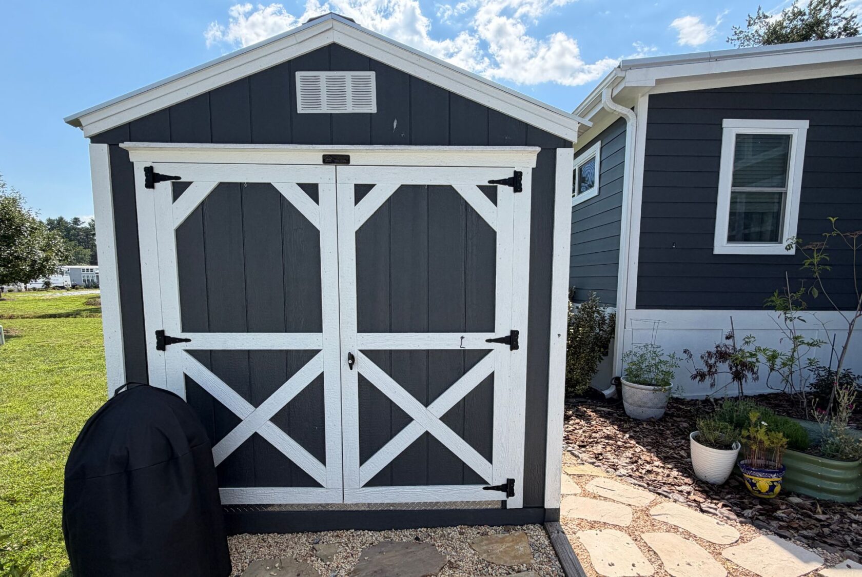 Gray and white garden shed at 103 Meandering Lane, with potted plants and a grill cover in the foreground.