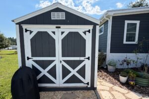 Gray and white garden shed at 103 Meandering Lane, with potted plants and a grill cover in the foreground.