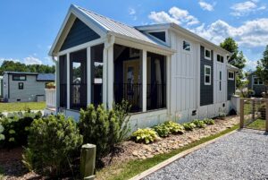A modern tiny house at 103 Meandering Lane with a screened porch, plants, and gravel path in a sunny, small community.
