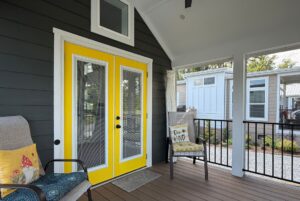 Covered porch at 103 Meandering Lane features two chairs, bright yellow double doors, and a “Be Kind” pillow.
