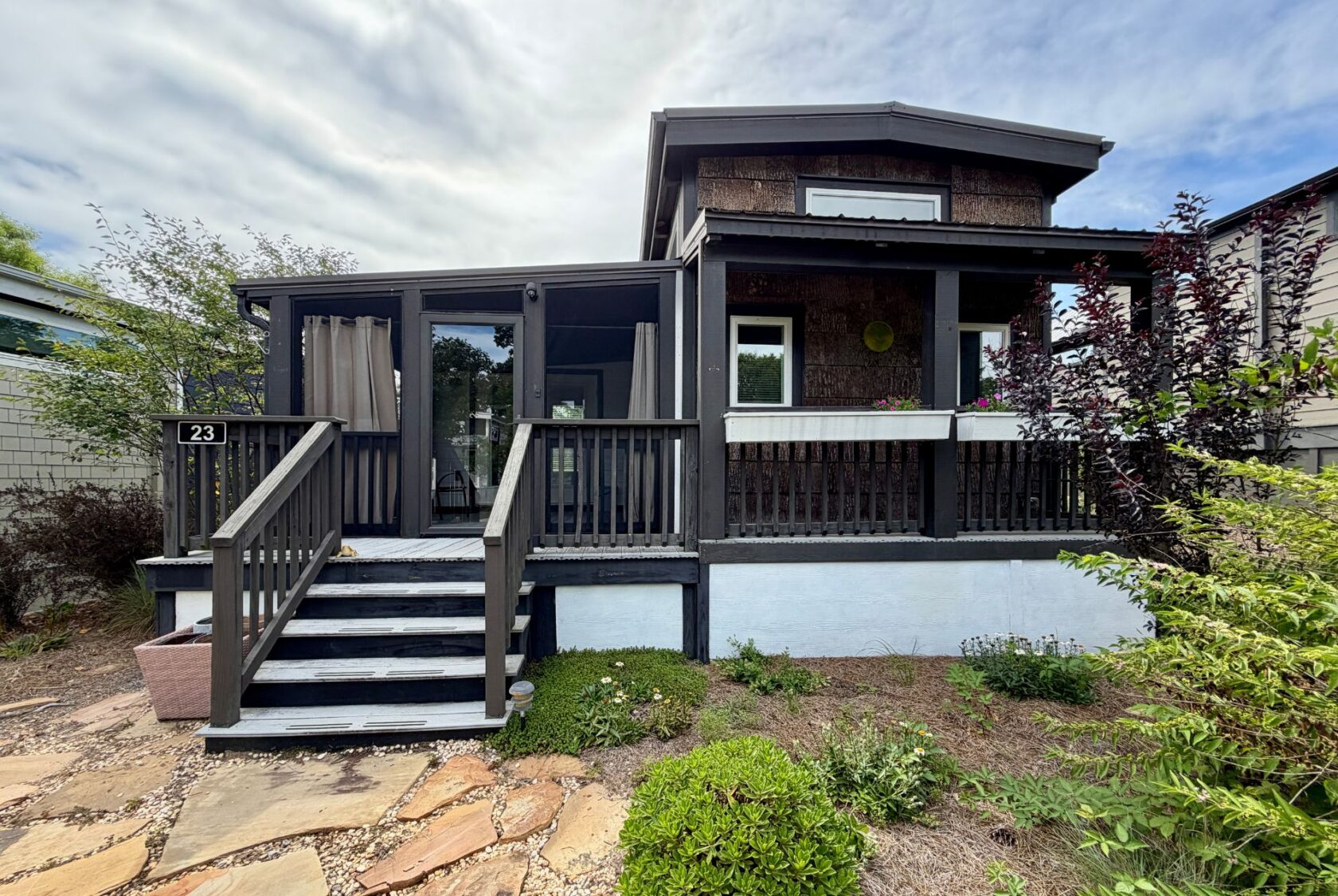 A small gray house on Highland Hill Lane, with a front porch, steps, and greenery under a partly cloudy sky.