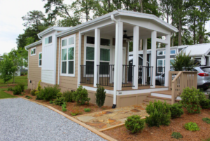 A small beige house with a front porch, white trim, and landscaping, surrounded by trees and a gravel driveway.