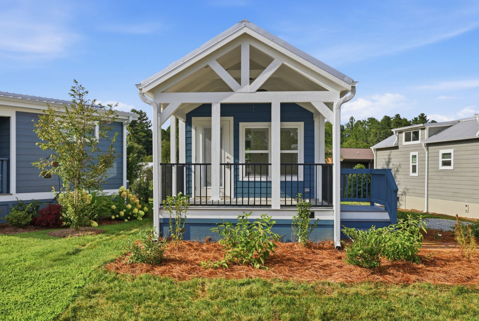 A small blue house with a white porch and railing, surrounded by green grass and shrubs under a blue sky.