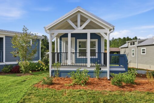 A small blue house with a white porch and railing, surrounded by green grass and shrubs under a blue sky.