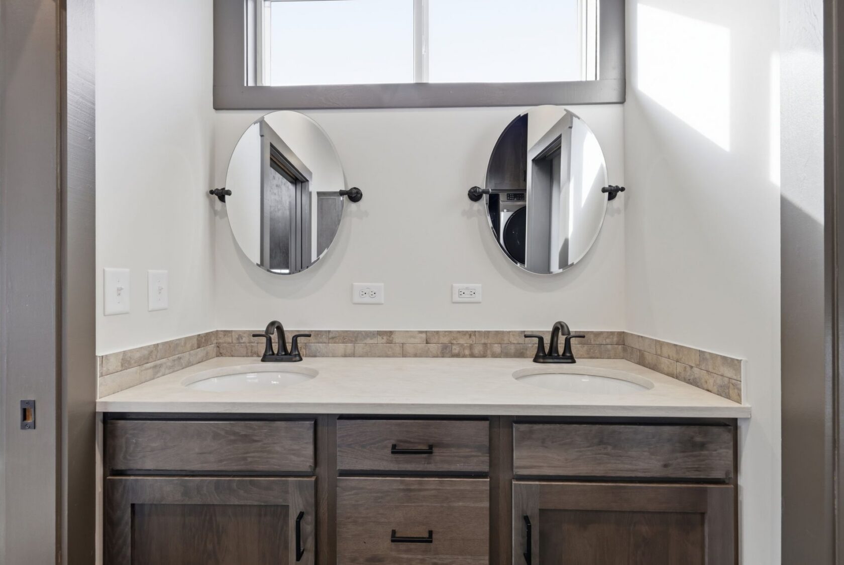 Double bathroom vanity with two round mirrors, two sinks, and wood cabinets under a bright window.