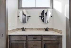 Double bathroom vanity with two round mirrors, two sinks, and wood cabinets under a bright window.