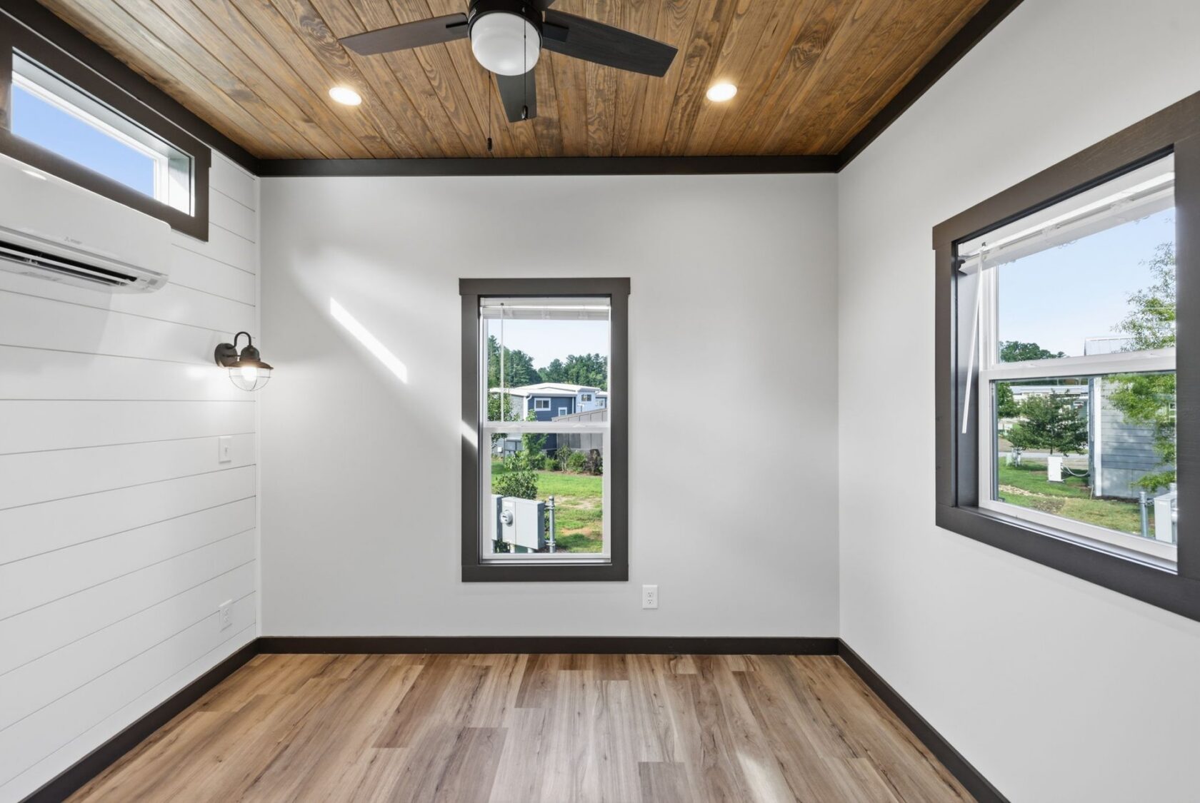 Bright empty room with wood ceiling, ceiling fan, large windows, wood floors, and white walls with dark trim.