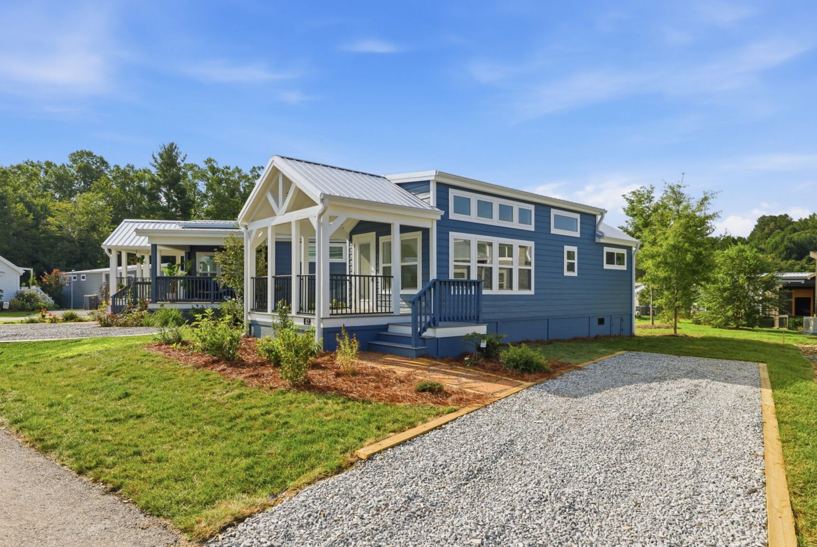 Blue tiny house with white trim, front porch, and gravel driveway on a sunny day.