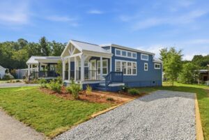 Blue tiny house with white trim, front porch, and gravel driveway on a sunny day.