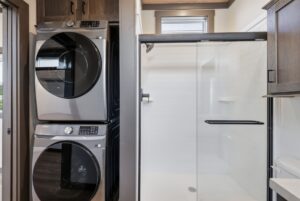 Stacked washer and dryer next to a glass-door shower in a modern bathroom with white tiles and wooden cabinets.