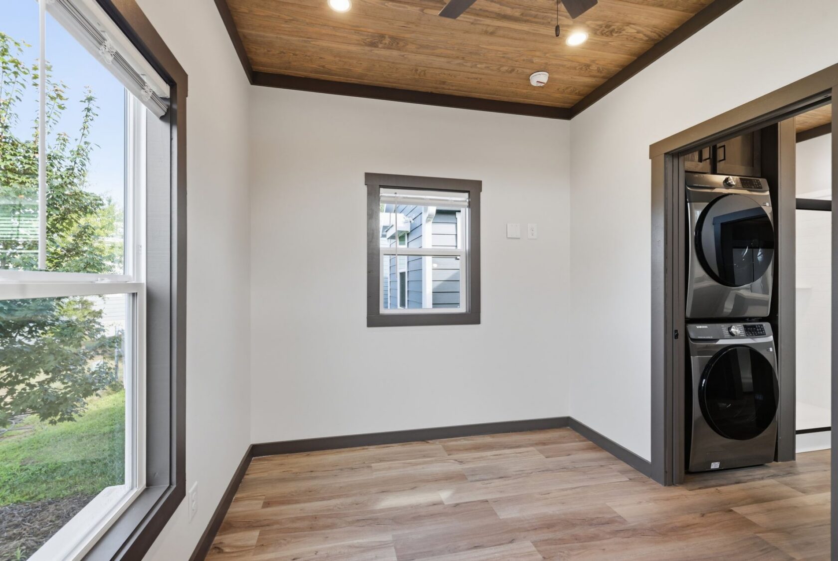 Small laundry room with wood floors, stacked washer and dryer, large windows, and wood ceiling with a ceiling fan.