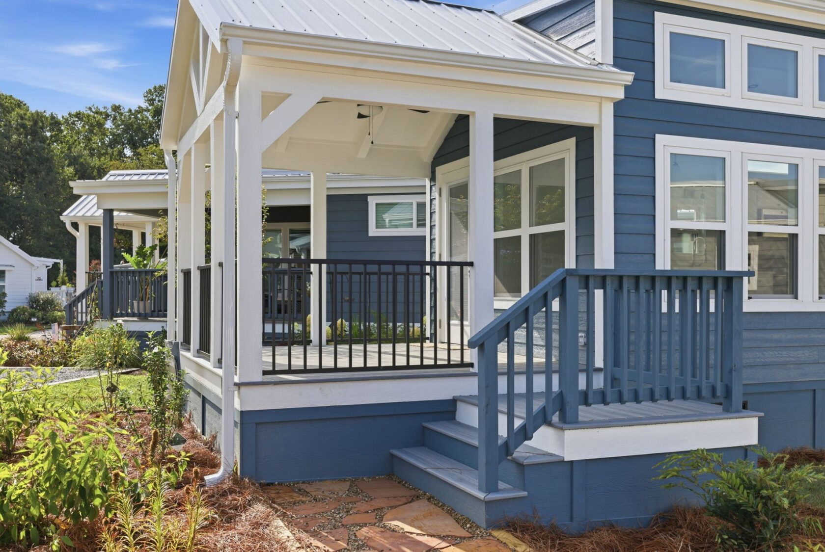 Blue house with white trim, front porch with steps and railing, surrounded by plants and a stone walkway.