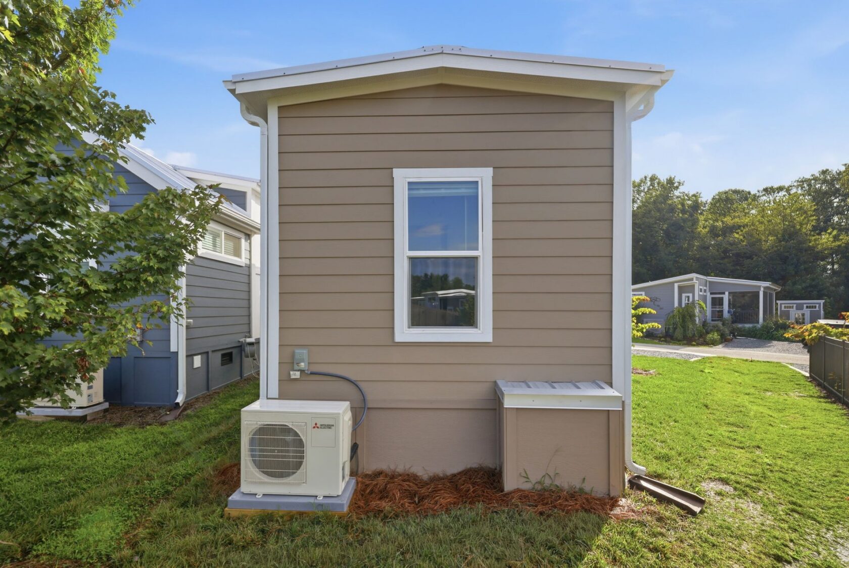 A small tan house with a single window, HVAC unit, and grass yard, seen from the back on a sunny day.
