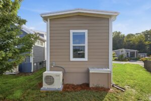 A small tan house with a single window, HVAC unit, and grass yard, seen from the back on a sunny day.
